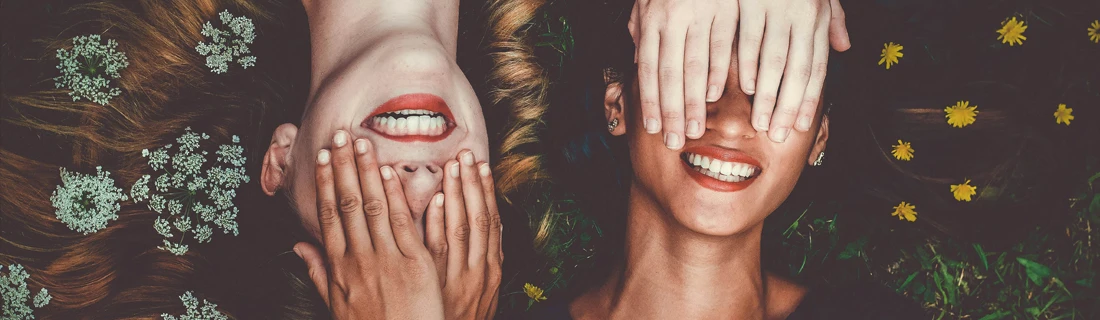 Two girls smiling with flowers in their hair. They are laying on he ground covering each other eyes.